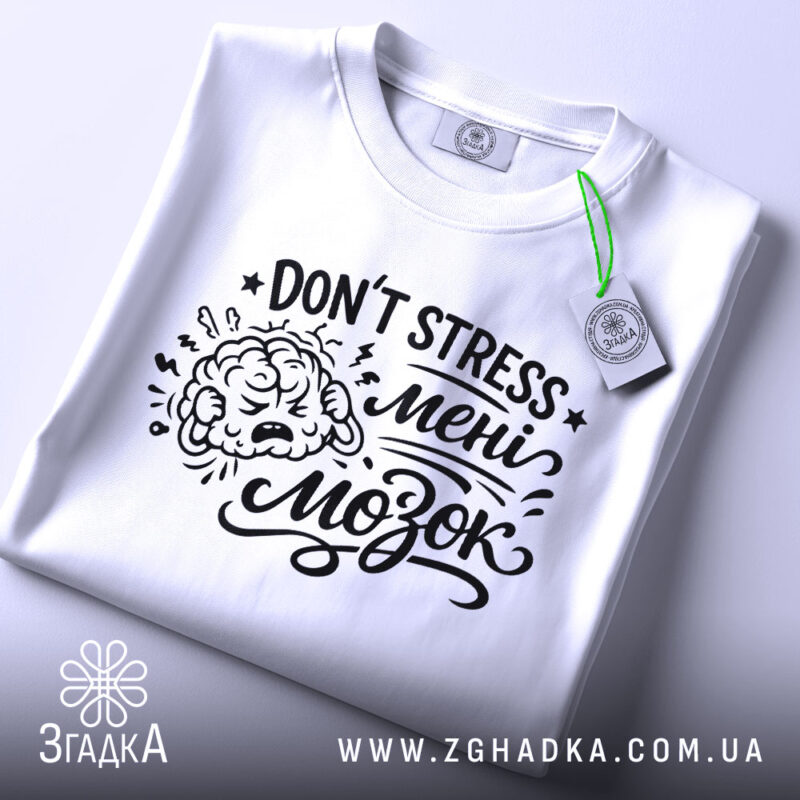Футболка Не стресуй мені мозок. Біла футболка, складена на поверхні, з видимим принтом чорного кольору. Текст 'DON'T STRESS мені мозок' та малюнок мозку. Фон світло-сірий, з елементами освітлення.