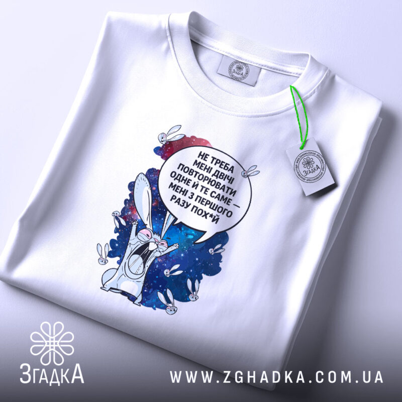 Футболка Не треба повторювати. Біла футболка, складена на столі, з принтом кролика на космічному фоні, текстом зверху. На футболці є етикетка з зеленим шнурком, фон світлий.