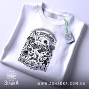 Футболка Не загубись, білого кольору, складена на поверхні, ракурс зверху, фон світло-фіолетовий, принт чорного кольору з українськими мотивами, на якому зображені люди, тварини та елементи природи, з етикеткою зеленого кольору.