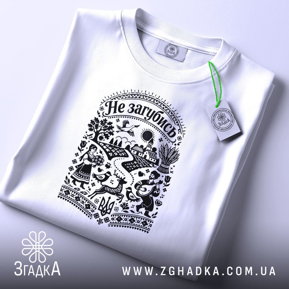 Футболка Не загубись, білого кольору, складена на поверхні, ракурс зверху, фон світло-фіолетовий, принт чорного кольору з українськими мотивами, на якому зображені люди, тварини та елементи природи, з етикеткою зеленого кольору.