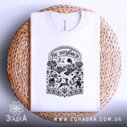 Футболка Не загубись, білого кольору, складена на кошику, ракурс зверху, фон світло-фіолетовий, принт чорного кольору з українськими мотивами, на якому зображені люди, тварини та елементи природи.