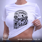 Футболка Не загубись, білого кольору, складена на поверхні, ракурс зверху, фон світло-фіолетовий, принт чорного кольору з українськими мотивами, на якому зображені люди, тварини та елементи природи, з етикеткою зеленого кольору.