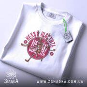 Футболка нехай щастить білого кольору, складена на поверхні, з видимим принтом скелета, що біжить, та написом 'Нехай щастить' у червоному кольорі. На футболці також є етикетка з зеленим шнурком.