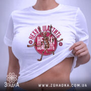 Футболка нехай щастить білого кольору, складена на поверхні, з видимим принтом скелета, що біжить, та написом 'Нехай щастить' у червоному кольорі. На футболці також є етикетка з зеленим шнурком.