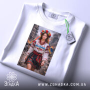 Футболка незламна патріотичний принт, біла, на плоскій поверхні, фон світло-фіолетовий, принт з дівчиною в національному вбранні, з квітами на голові та написом 'Незламна', з етикеткою на комірці.