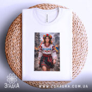 Футболка незламна патріотичний принт, біла, на плоскій поверхні, фон світло-фіолетовий, принт з дівчиною в національному вбранні, з квітами на голові та написом 'Незламна', розташована на плетеній основі.