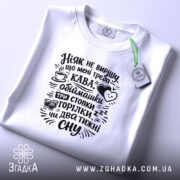 Футболка ніяк не вирішу що треба, біла, на плоскій поверхні, фон світло-сірий, принт чорний з текстом про каву та обіймашки, з етикеткою.