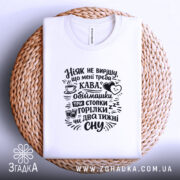 Футболка ніяк не вирішу що треба, біла, на плоскій поверхні, фон плетений, принт чорний з текстом про каву та обіймашки.