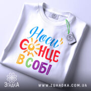 Футболка носи сонце в собі, біла, складена на поверхні, ракурс зверху, фон світлий, принт з написом 'Носи сонце в собі' у яскравих кольорах: червоний, жовтий, синій, зелений та фіолетовий, з елементами сонця, на ярлику видно логотип.