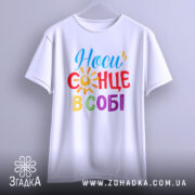 Футболка носи сонце в собі, біла, підвішена на вішалці, ракурс спереду, фон світлий, принт з написом 'Носи сонце в собі' у яскравих кольорах: червоний, жовтий, синій, зелений та фіолетовий, з елементами сонця.