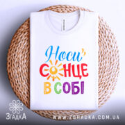 Футболка носи сонце в собі, біла, складена на плетеній кошику, ракурс зверху, фон світлий, принт з написом 'Носи сонце в собі' у яскравих кольорах: червоний, жовтий, синій, зелений та фіолетовий, з елементами сонця.