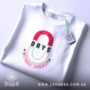 Футболка Нуль днів без сарказму, біла, на столі, з етикеткою, фон світло-сірий, принт з червоними і чорними літерами, напис '0 DAYS WITHOUT SARCASM' з зірочками.