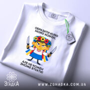 Футболка образити може кожен. Біла футболка, зображена зверху, на фоні світло-фіолетового кольору. Принт з ілюстрацією дівчинки в українському вбранні, тримає ножі, текст чорного кольору розташований зверху і знизу.