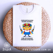 Футболка образити може кожен. Біла футболка, зображена зверху на плетеному кошику, фон світлий. Принт з ілюстрацією дівчинки в українському вбранні, тримає ножі, текст чорного кольору розташований зверху і знизу.