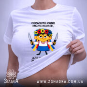 Футболка образити може кожен. Біла футболка, зображена зверху, на фоні світло-фіолетового кольору. Принт з ілюстрацією дівчинки в українському вбранні, тримає ножі, текст чорного кольору розташований зверху і знизу.
