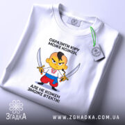 Футболка образити може кожен. На фото зображена біла футболка, яка лежить на поверхні. Принт з мультяшним персонажем, який тримає два мечі, розташований в центрі. На футболці видно етикетку з логотипом бренду, фон нейтральний, що акцентує увагу на футболці.