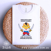 Футболка образити може кожен. На фото зображена біла футболка, яка лежить на плетеній кошику. Принт з мультяшним персонажем, який тримає два мечі, розташований в центрі. Фон нейтральний, що підкреслює текстуру кошика.