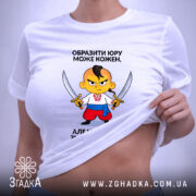 Футболка образити може кожен. На фото зображена біла футболка, яка лежить на поверхні. Принт з мультяшним персонажем, який тримає два мечі, розташований в центрі. На футболці видно етикетку з логотипом бренду, фон нейтральний, що акцентує увагу на футболці.