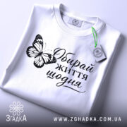 Футболка обирай життя щодня, біла, на столі, складена, з принтом метелика та написом, фон світло-сірий, деталі принта яскраві.