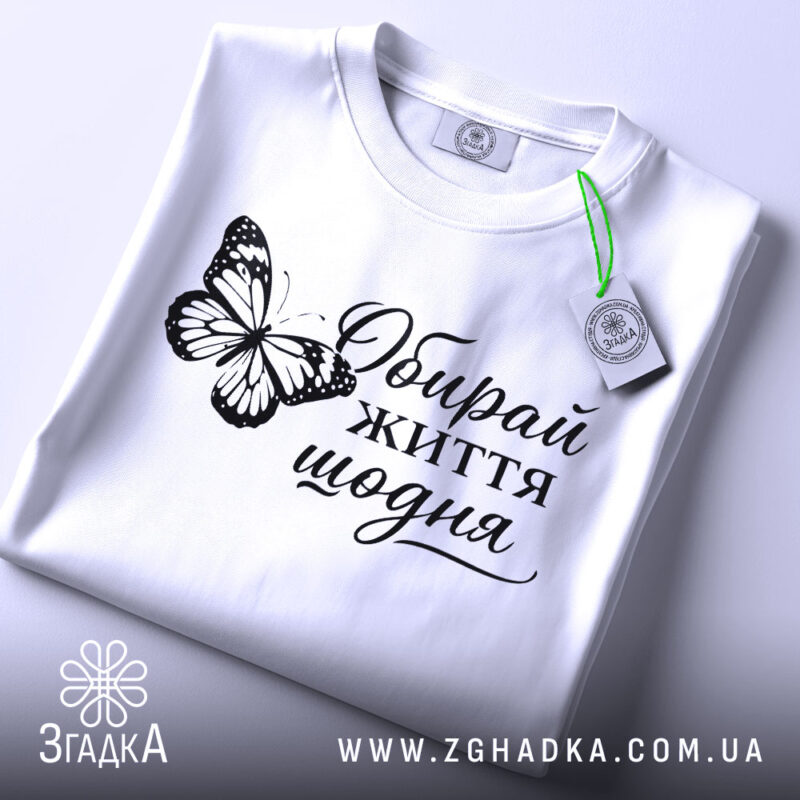 Футболка обирай життя щодня, біла, на столі, складена, з принтом метелика та написом, фон світло-сірий, деталі принта яскраві.
