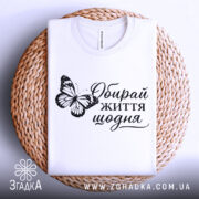 Футболка обирай життя щодня, біла, на плетеній кошику, складена, з принтом метелика та написом, фон світло-блакитний, деталі принта чіткі.