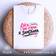 Футболка ой я не пʼяна, на фото зображена складена біла футболка на плетеній кошику, принт яскравий, текст виконаний у чорному та рожевому кольорах, фон світло-сірий.