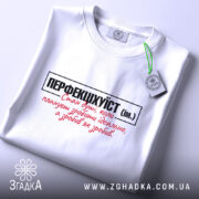 Футболка перфекціхуїст білого кольору, складена на поверхні, з видимим етикеткою зеленого кольору. Принт на футболці чорного та червоного кольорів, чітко видимий на фоні. Освітлення м'яке, створює спокійну атмосферу.