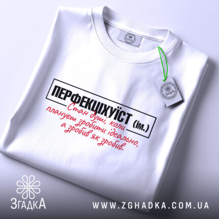 Футболка перфекціхуїст білого кольору, складена на поверхні, з видимим етикеткою зеленого кольору. Принт на футболці чорного та червоного кольорів, чітко видимий на фоні. Освітлення м'яке, створює спокійну атмосферу.