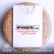 Футболка перфекціхуїст білого кольору, складена на плетеній кошику, з чітким принтом чорного та червоного кольорів. Освітлення м'яке, фон нейтральний, що підкреслює текст на футболці.