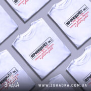 Футболка перфекціхуїст білого кольору, зображена на чоловікові, який позує з легким усмішкою. Принт на футболці чорного та червоного кольорів, розташований по центру, з текстом, що говорить про ідеальність. Фон м'який, світлий, що підкреслює яскравість футболки.