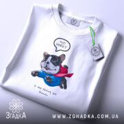 Футболка Пес супергерой, біла, на столі, фон світло-сірий, принт з зображенням собаки в синьому костюмі та червоному плащі, текст "NO WORRY" над собакою і "I am saving you Human" під ним, з етикеткою на рукаві.