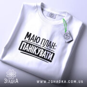 Футболка План панікувати, біла, складена на поверхні, з верхнього ракурсу, фон світло-сірий, принт чорного кольору з текстом 'МАЮ ПЛАН: ПАНІКУВАТИ', з етикеткою зеленого кольору.
