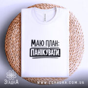 Футболка План панікувати, біла, складена на плетеній тарілці, з верхнього ракурсу, фон світло-сірий, принт чорного кольору з текстом 'МАЮ ПЛАН: ПАНІКУВАТИ'.