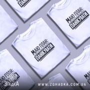 Футболка План панікувати, біла, на моделі, з легким ракурсом спереду, фон світло-фіолетовий, принт чорного кольору з текстом 'МАЮ ПЛАН: ПАНІКУВАТИ'.