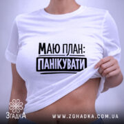 Футболка План панікувати, біла, складена на поверхні, з верхнього ракурсу, фон світло-сірий, принт чорного кольору з текстом 'МАЮ ПЛАН: ПАНІКУВАТИ', з етикеткою зеленого кольору.
