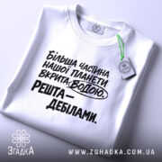 Футболка планета вкрита водою, біла футболка на столі, ракурс зверху, фон світлий, принт чорного кольору з текстом: 'Більша частина нашої планети вкрита водою. Решта – дебілами.'