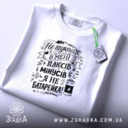 Футболка плюси мінуси: складена біла футболка на світлому фоні, з видимим принтом чорного кольору, на якому написано текст, з зеленим ярликом.
