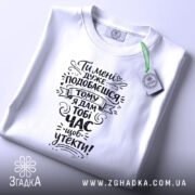 Футболка подобаєшся але біжи швидше. Біла футболка, складена на поверхні, зображена зверху. На фоні світло-фіолетовий колір. Принт чорного кольору з текстом, оформлений у стилі рукописного шрифту, а також видно етикетку з зеленим шнурком.