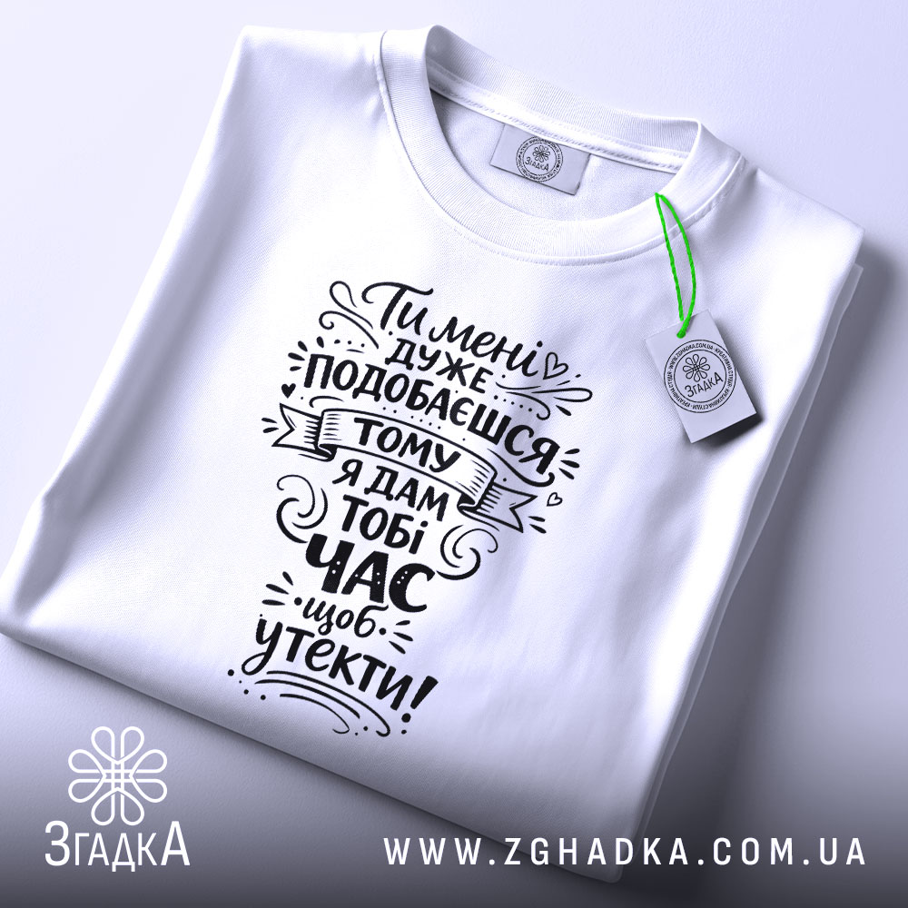 Футболка подобаєшся але біжи швидше. Біла футболка, складена на поверхні, зображена зверху. На фоні світло-фіолетовий колір. Принт чорного кольору з текстом, оформлений у стилі рукописного шрифту, а також видно етикетку з зеленим шнурком.