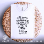 Футболка подобаєшся але біжи швидше. Біла футболка, складена на плетеній кошику, зображена зверху. На фоні світло-фіолетовий колір. Принт чорного кольору з текстом, оформлений у стилі рукописного шрифту.