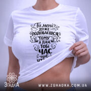 Футболка подобаєшся але біжи швидше. Біла футболка, складена на поверхні, зображена зверху. На фоні світло-фіолетовий колір. Принт чорного кольору з текстом, оформлений у стилі рукописного шрифту, а також видно етикетку з зеленим шнурком.