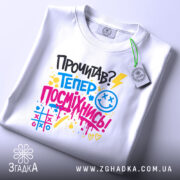 Футболка посміхнись. Біла футболка, зображена зверху, на світлому фоні. Принт з текстом 'Прочитав? Тепер посміхнись!' виконаний у яскравих кольорах, з елементами графіки, та з етикеткою на рукаві.