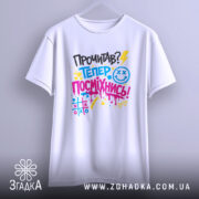 Футболка посміхнись. Біла футболка, підвішена на вішалці, фон світло-сірий. Принт з текстом 'Прочитав? Тепер посміхнись!' у яскравих кольорах, з елементами графіки, добре видимий.