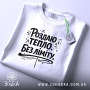 Футболка роздаю тепло без ліміту, біла, з верхнього ракурсу, на плоскій поверхні, фон світло-сірий, принт чорного кольору з написом 'Роздаю тепло без ліміту' та елементами дизайну, з етикеткою зеленого кольору.