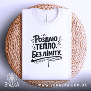 Футболка роздаю тепло без ліміту, біла, з верхнього ракурсу, на плоскій поверхні, фон світло-сірий, принт чорного кольору з написом 'Роздаю тепло без ліміту' та елементами дизайну, розташована на плетеній основі.