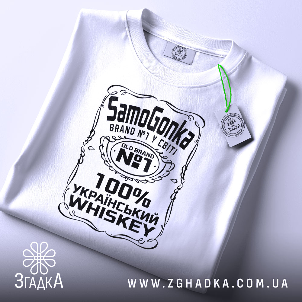 Футболка самогонка, біла, на столі, з верхнього ракурсу, фон світло-сірий, принт чорного кольору з написом 'SamoGonka', '100% УКРАЇНСЬКИЙ WHISKEY' та етикеткою з зеленим шнурком.