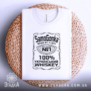 Футболка самогонка, біла, на столі, з верхнього ракурсу, фон світло-сірий, принт чорного кольору з написом 'SamoGonka', '100% УКРАЇНСЬКИЙ WHISKEY', розташована на плетеній основі.