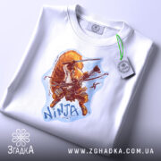 Футболка самурайська білка, біла, на столі, ракурс зверху, фон світло-фіолетовий, принт з зображенням білки-самурая, на футболці є етикетка з логотипом.