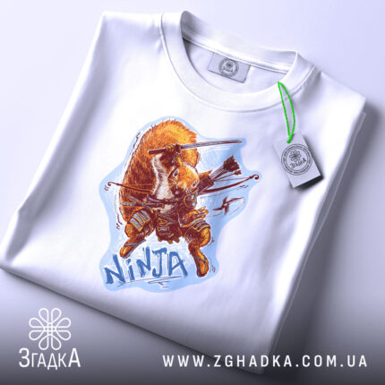 Футболка самурайська білка, біла, на столі, ракурс зверху, фон світло-фіолетовий, принт з зображенням білки-самурая, на футболці є етикетка з логотипом.