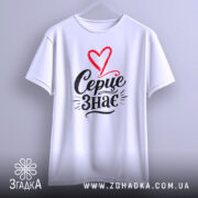 Футболка Серце знає, біла, підвішена на вішалці, фон світло-сірий, принт з червоним серцем і чорним написом 'Серце знає'.